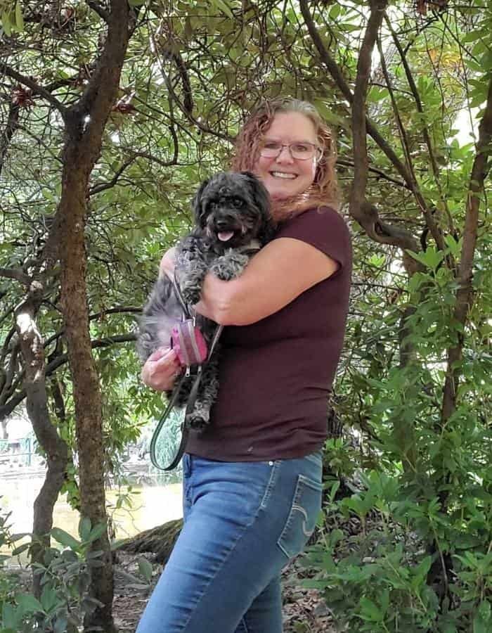 Sherry holding Shelby, a black Yorkiepoo, in a forest of green trees