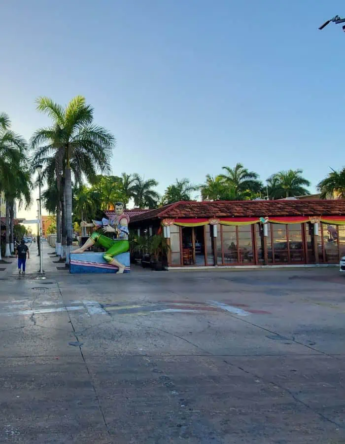 A street scene shows a building with a red roof and glass walls surrounded by palm trees. A decorative fountain with a green dolphin sculpture stands nearby under a clear blue sky.