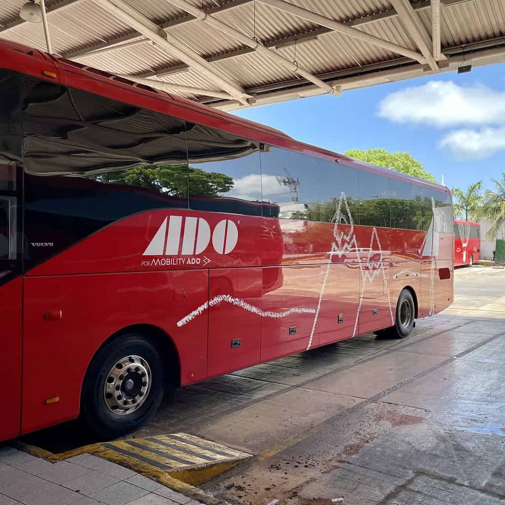 A red ADO passenger bus parked at a bus terminal ready to take people from Tulum to Playa del Carmen, with the company's white logo on the side, under a partially shaded area with trees and a clear blue sky in the background