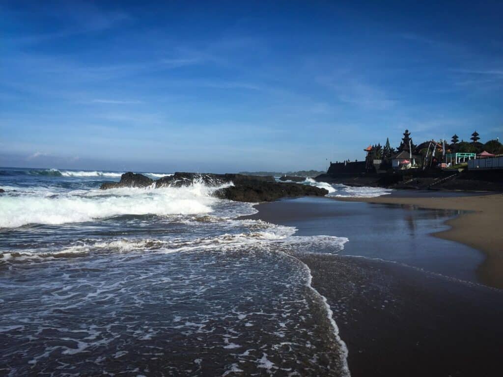 Gently crashing waves roll onto a dark sandy beach under a clear blue sky, with a small seaside village visible in the distance along the shoreline. The scene captures a peaceful coastal atmosphere with reflective wet sand and rocky outcrops.