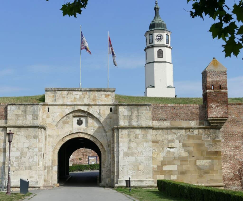 Stone entrance gate of Belgrade Fortress with two flags flying above, leading to a historic path within the walls. In the background, the tall white clock tower rises against a clear blue sky, framed by patches of green grass and leafy trees.