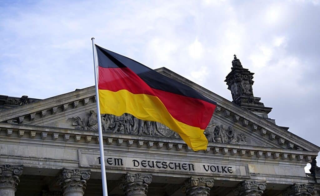 German flag waves prominently in front of the Reichstag building in Berlin, with its neoclassical façade and the inscription "DEM DEUTSCHEN VOLKE" (To the German People) visible beneath the pediment. The cloudy sky adds a dramatic backdrop to this symbol of German government and history.