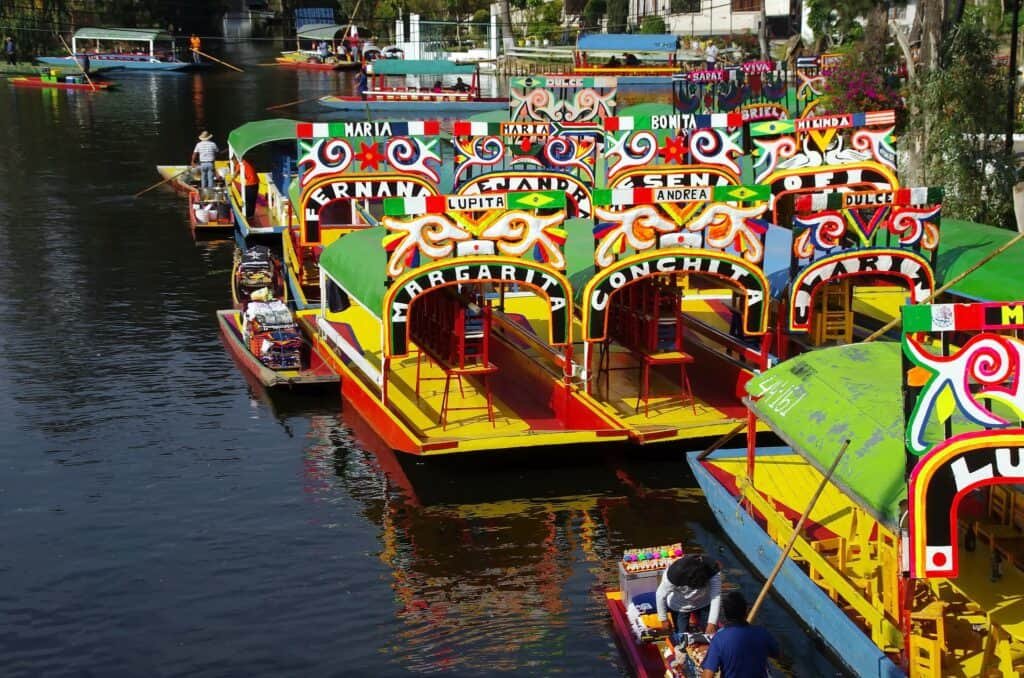 Colorful wooden boats with vibrant painted arches float closely together on a calm canal. Each boat has a name displayed on the arch, surrounded by decorative swirls and bright patterns.