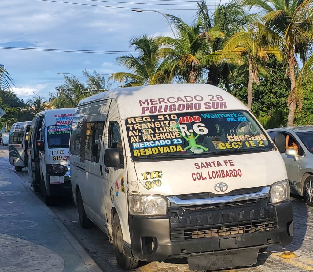 White public minibus in Cancún with colorful route information painted across the windshield, including destinations like Mercado 28, Polígono Sur, and Santa Fe. The bus is parked on a sunny street lined with palm trees, with other similar minibuses behind it.