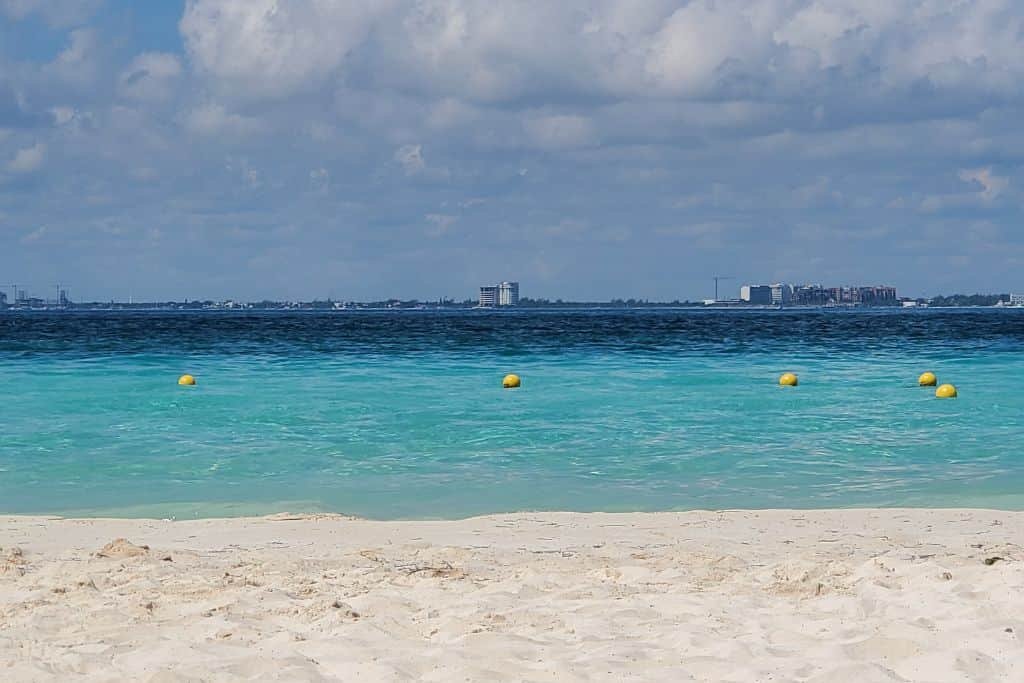 A serene beach with soft, light-colored sand in the foreground, leading to the calm turquoise waters of the ocean. Yellow buoys float on the water, creating a sense of a designated swimming area. In the distance, across the water, the skyline of Cancun is visible under a partly cloudy sky.