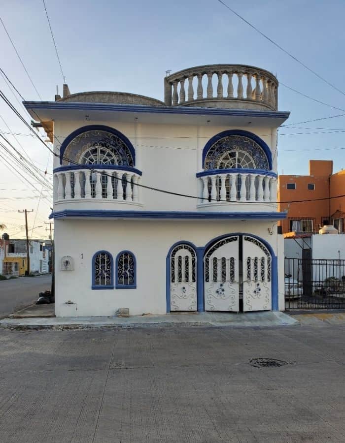 The image shows a two-story white house with blue trim and decorative arched windows. The upper floor features a curved balcony with white balusters and a round rooftop terrace. The ground level has two arched doors and a window framed in blue tile.