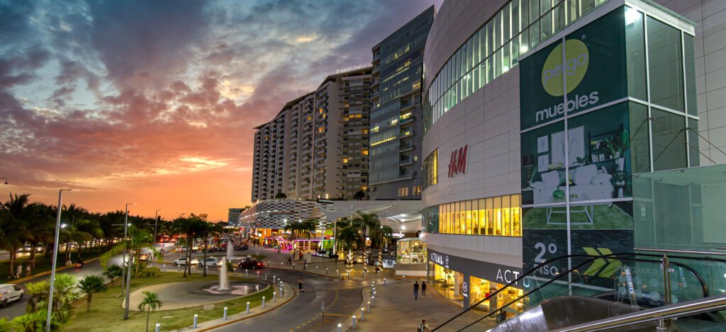 A modern shopping mall in Cancun during sunset, with a multi-story building featuring stores like H&M and a large advertisement for "Pego Muebles." The street is lined with palm trees, cars, and a circular fountain in the foreground, while the sky is filled with dramatic, colorful hues from the setting sun.