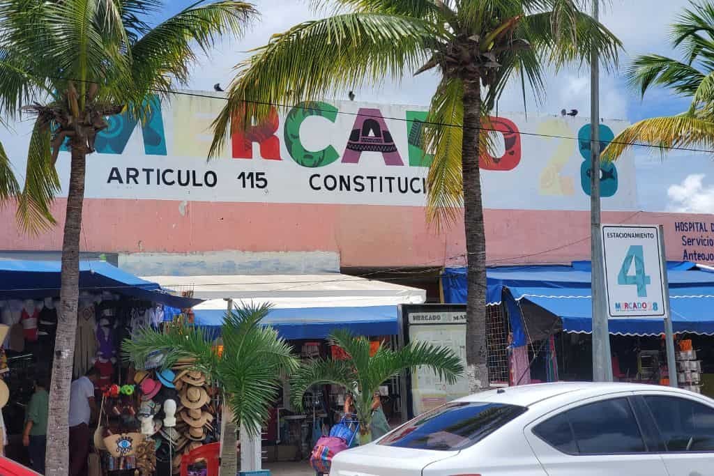 The entrance of "Mercado 28" in Cancun, a market building with a colorful sign that reads "MERCADO 28 ARTICULO 115 CONSTITUCION." Palm trees line the front of the market, and several stalls with hats and other goods are visible under blue awnings. A car is parked in the foreground, highlighting the busy, bustling atmosphere of the market area.