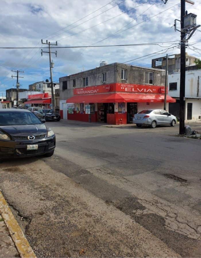 Street view of a local neighborhood in Cancún featuring a corner building with a bright red awning labeled “Abarrotes Silvia,” a small convenience store. Cars are parked along the cracked pavement, and overhead power lines stretch across the partly cloudy sky.
