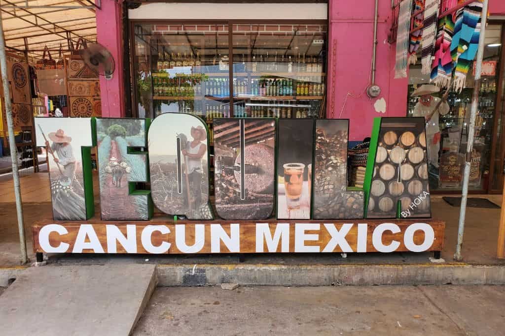 Large decorative sign that spells “TEQUILA” with images of agave plants, barrels, and tequila production inside the letters, sitting above the words “CANCUN MEXICO” in bold white and orange letters. The sign is displayed outside a colorful storefront with souvenirs visible in the background.