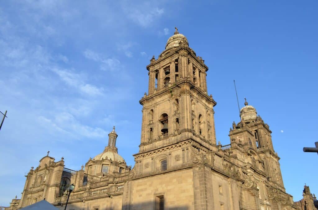 A large stone cathedral with ornate towers and domes rises against a bright blue sky. The detailed architecture features columns and carvings, with warm beige stone that catches the sunlight.