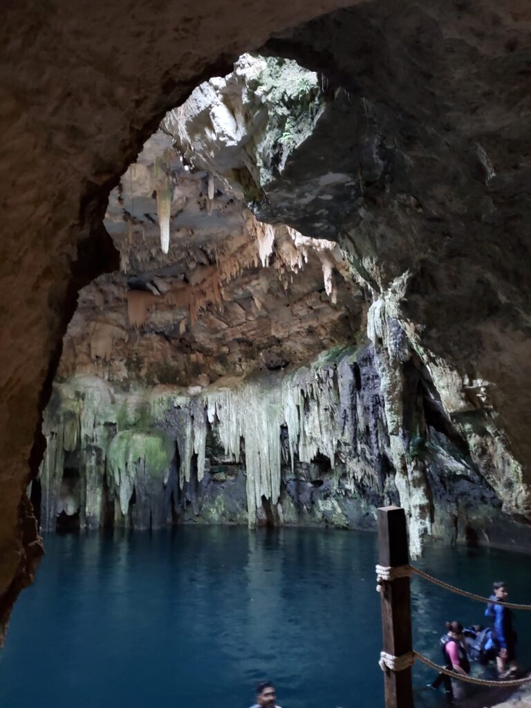 The inside of a cenote, a natural underground cave filled with water. Stalactites hang from the rocky ceiling, while light filters in through an opening, illuminating the textured rock formations and the calm, dark blue water below. A group of people stands at the edge of the cenote, adding a sense of scale and exploration to this mystical natural setting.