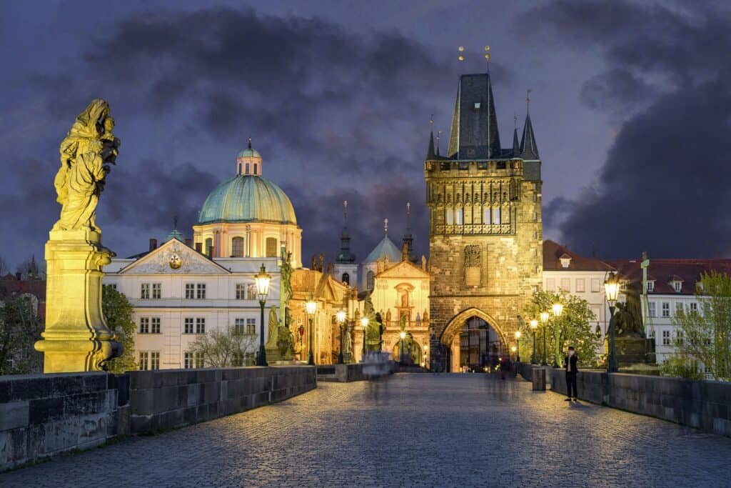 Evening view of Prague’s Charles Bridge illuminated by warm lights, featuring statues along the cobblestone path and the iconic Old Town Bridge Tower rising in the background. The dome of St. Francis of Assisi Church and historic buildings frame the moody, cloud-filled sky.