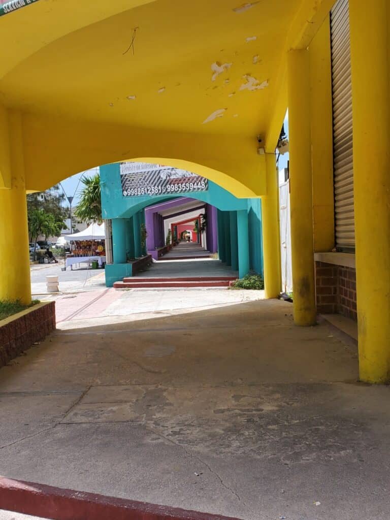 Colorful walkway in Cancún with a series of archways painted in bright yellow, turquoise, purple, and red, creating a tunnel-like effect. Sunlight casts shadows on the pavement, and trees and buildings can be seen beyond the arches at the end of the path.