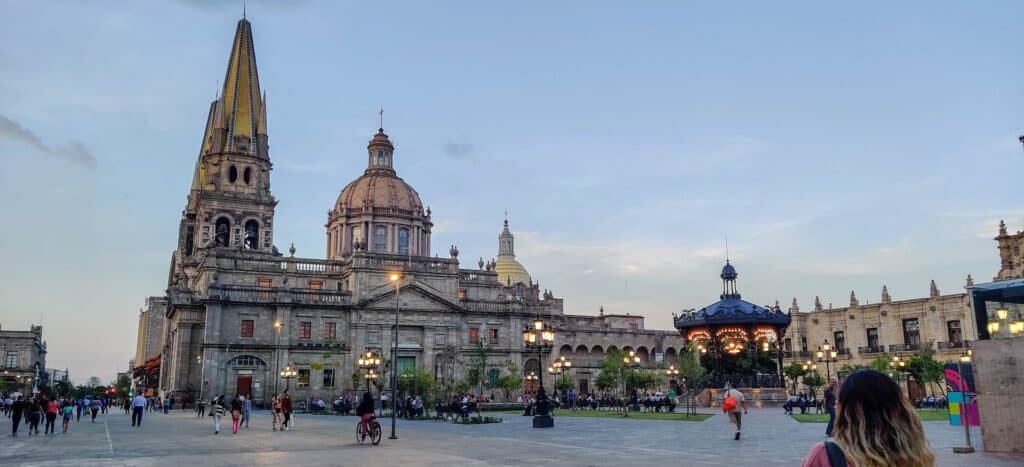 A historic cathedral with a tall pointed tower and domed roof overlooks a large public square. People stroll near a central gazebo and decorative lights as the sky begins to darken at dusk.
