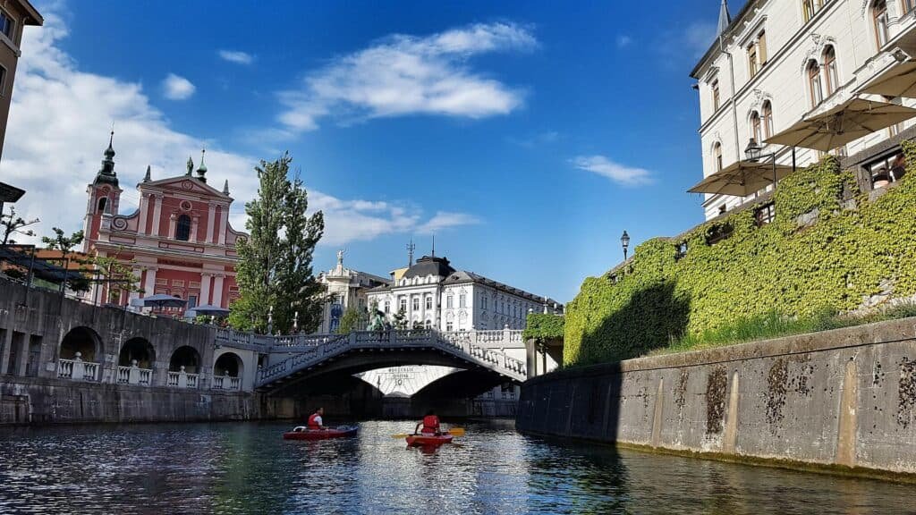 Scenic view of the Ljubljanica River in Ljubljana, Slovenia, with kayakers paddling beneath a historic stone bridge. The scene is framed by ivy-covered walls, elegant buildings, and the iconic pink Franciscan Church of the Annunciation under a bright blue sky.