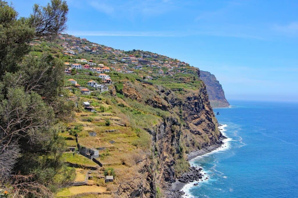 Steep coastal cliffs of Madeira Island rise above the deep blue Atlantic Ocean, dotted with terraced fields and scattered homes. The rugged landscape is lush with greenery under a clear, sunny sky, showcasing the island's dramatic natural beauty.