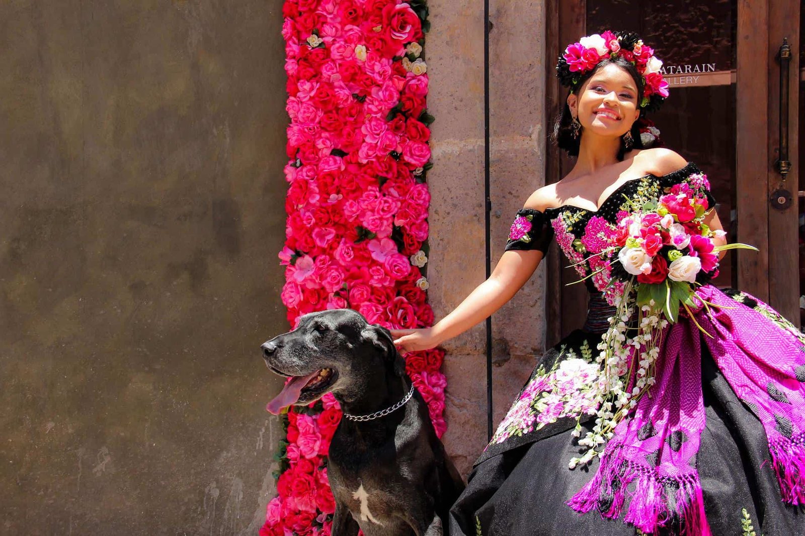 A smiling young woman in a vibrant black and purple floral gown stands beside a large black dog, holding a colorful bouquet. She wears a flower crown and stands in front of a wall adorned with bright pink flowers, creating a festive and elegant scene.