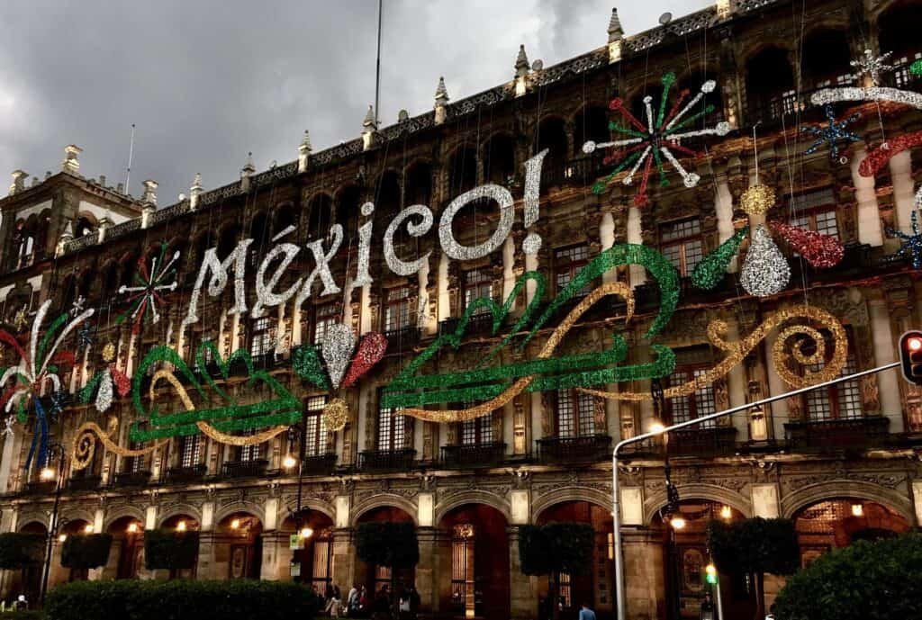 Historic building in Mexico City decorated with vibrant lights spelling out “¡México!” along with festive designs of fireworks, eagles, and the national colors green, white, and red. The illuminated display celebrates Mexican pride, likely for Independence Day, under a dramatic cloudy sky.