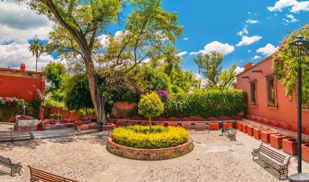 A charming courtyard garden with cobblestone paths features a circular planter filled with greenery and a small tree in the center. Red walls, benches, and tall trees surround the space under a bright blue sky with scattered clouds.