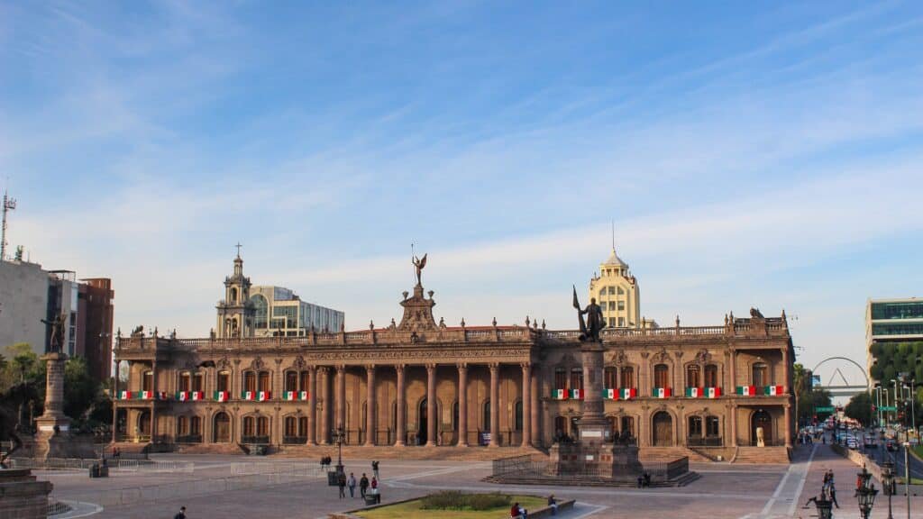 A grand neoclassical government building with tall columns and statues sits at the center of a spacious public plaza. People walk across the open square under a bright blue sky.