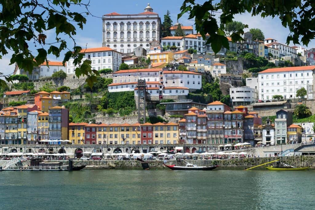Colorful hillside buildings line the Douro River in Porto, Portugal, with traditional boats docked along the waterfront. The scene features vibrant facades, red-tiled roofs, and the prominent white Monastery of Serra do Pilar perched at the top.