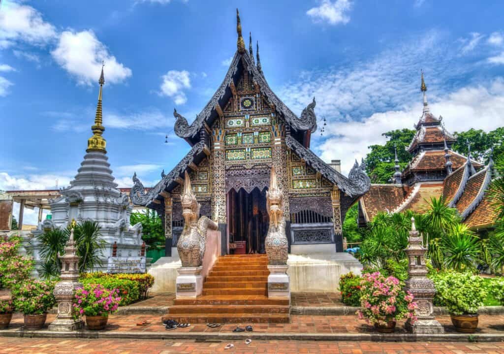 Ornate Thai temple with intricate wooden carvings and a steep, pointed roof, flanked by stone guardians and surrounded by lush greenery and manicured plants. A bright blue sky with scattered clouds adds vibrancy to the richly detailed structure and adjacent white and gold stupa.