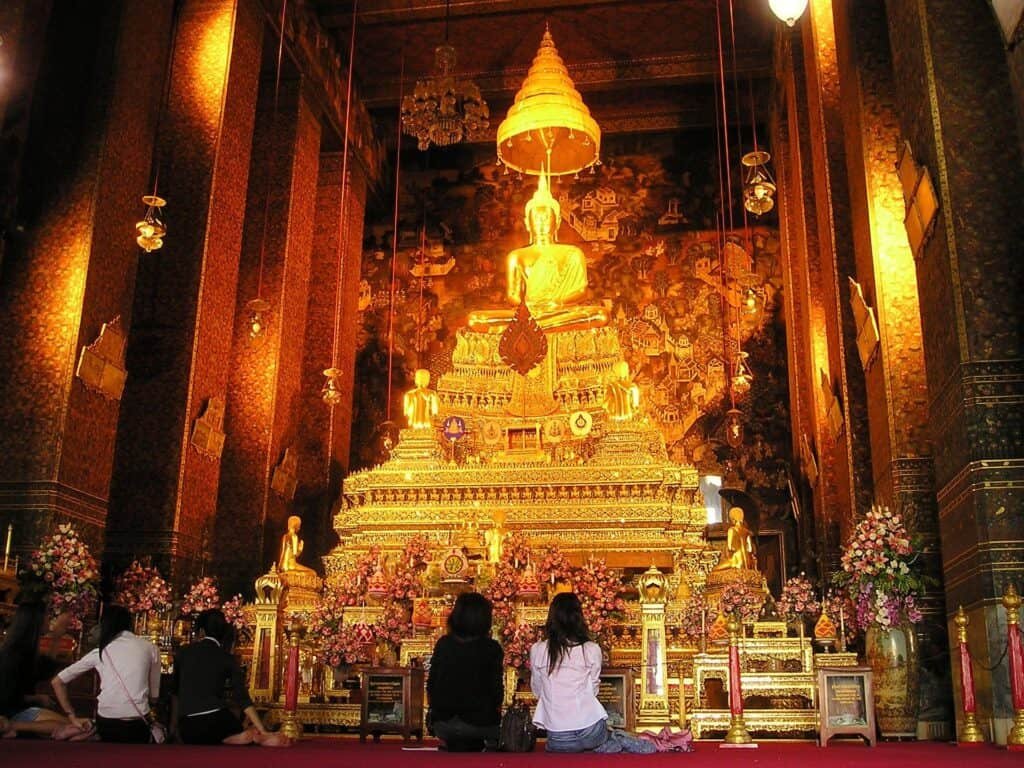 Interior of a Thai temple glowing with golden light, where visitors kneel before an ornate golden Buddha shrine adorned with floral offerings, intricate carvings, and a tiered umbrella above the statue. The rich red and gold décor creates a reverent and majestic atmosphere.