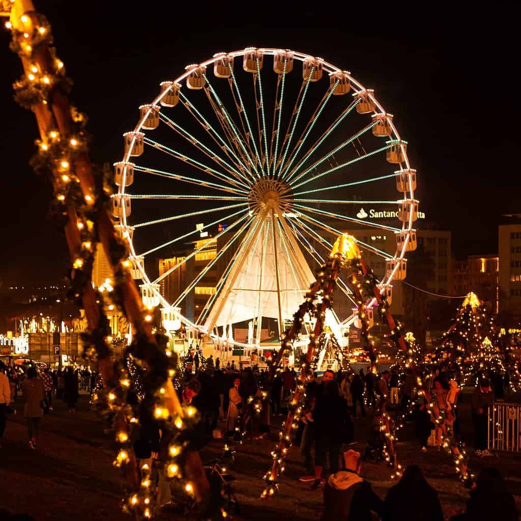 A glowing Ferris wheel lights up the night sky during Christmas in Lisbon, surrounded by festive trees wrapped in string lights. Crowds of people stroll through the illuminated holiday market, creating a warm, celebratory atmosphere.