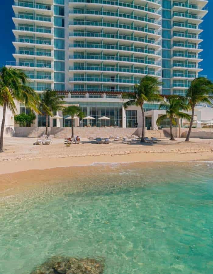 A modern beachfront high-rise with glass balconies overlooks golden sand and clear turquoise water. Palm trees line the shore, and lounge chairs are arranged near the water’s edge under a bright blue sky.