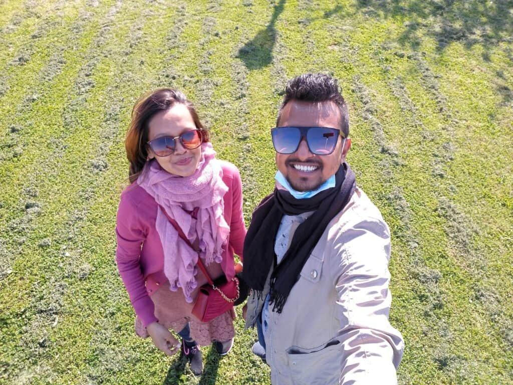 Two people wearing sunglasses and scarves pose for a video on a grassy field. Bright sunlight casts shadows, suggesting a cool but sunny day outdoors.