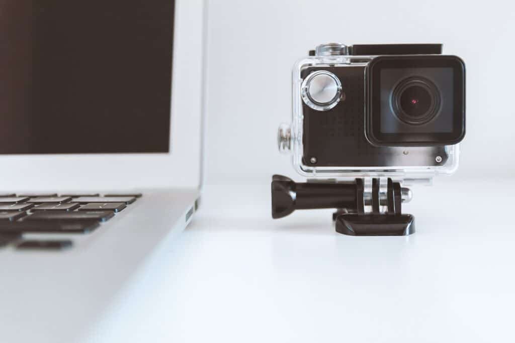 Close-up of a laptop next to an action camera on a white desk. The minimalist setup suggests content creation or travel vlogging preparation.