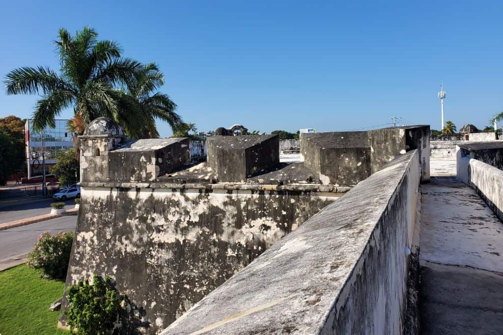 View from atop a weathered stone fort wall in Campeche, Mexico, featuring aged battlements and a palm tree-lined street below. The clear blue sky and bright sunlight highlight the historic structure's texture and surroundings.