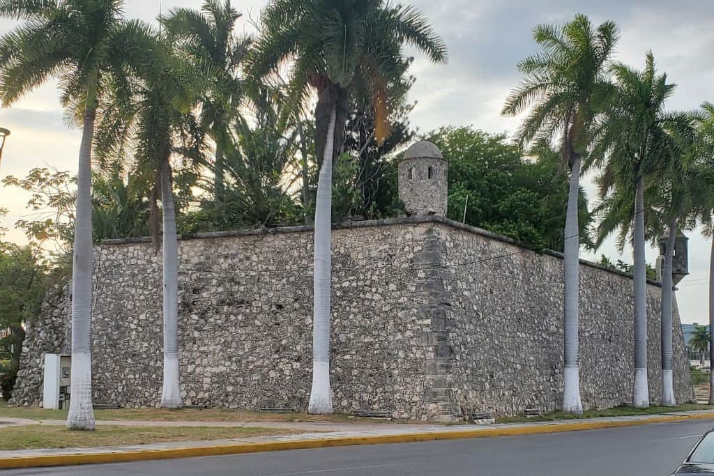 Stone fortification in Campeche, Mexico, surrounded by tall palm trees and situated at a street corner. The thick walls feature a small watchtower at the corner, hinting at the city's colonial military history.