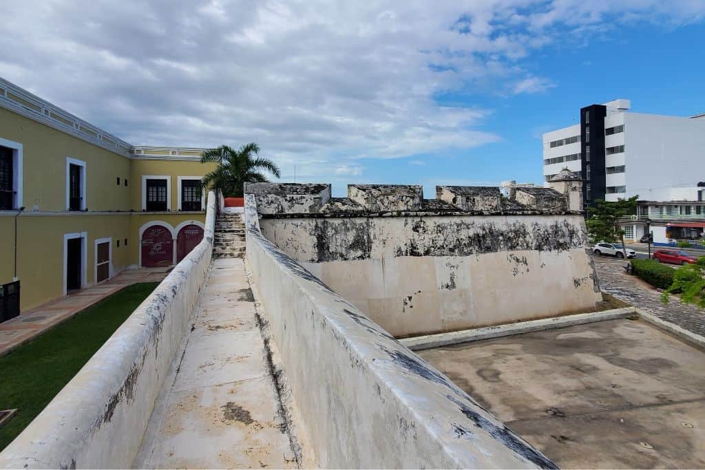 Stone walkway along a historic fortified wall in Campeche, Mexico, with weathered battlements and a view of colonial-style buildings on the left and modern structures on the right. The scene is set under a partly cloudy sky.