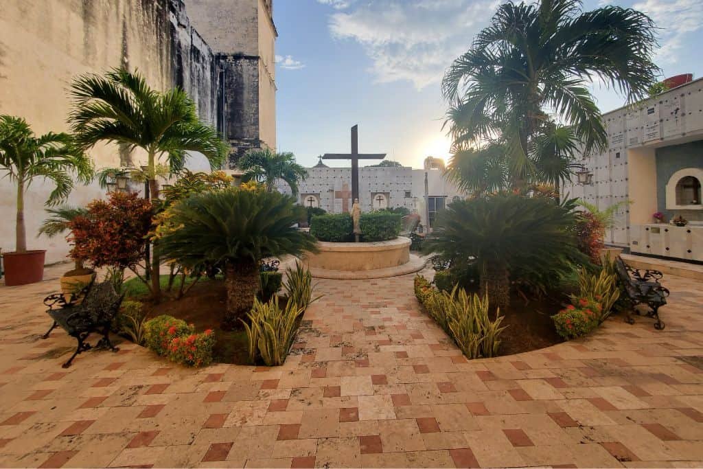 Peaceful courtyard in Campeche with a large wooden cross at the center, surrounded by tropical plants and palm trees. The scene is warmly lit by the setting sun, casting a golden glow across the patterned tile ground.