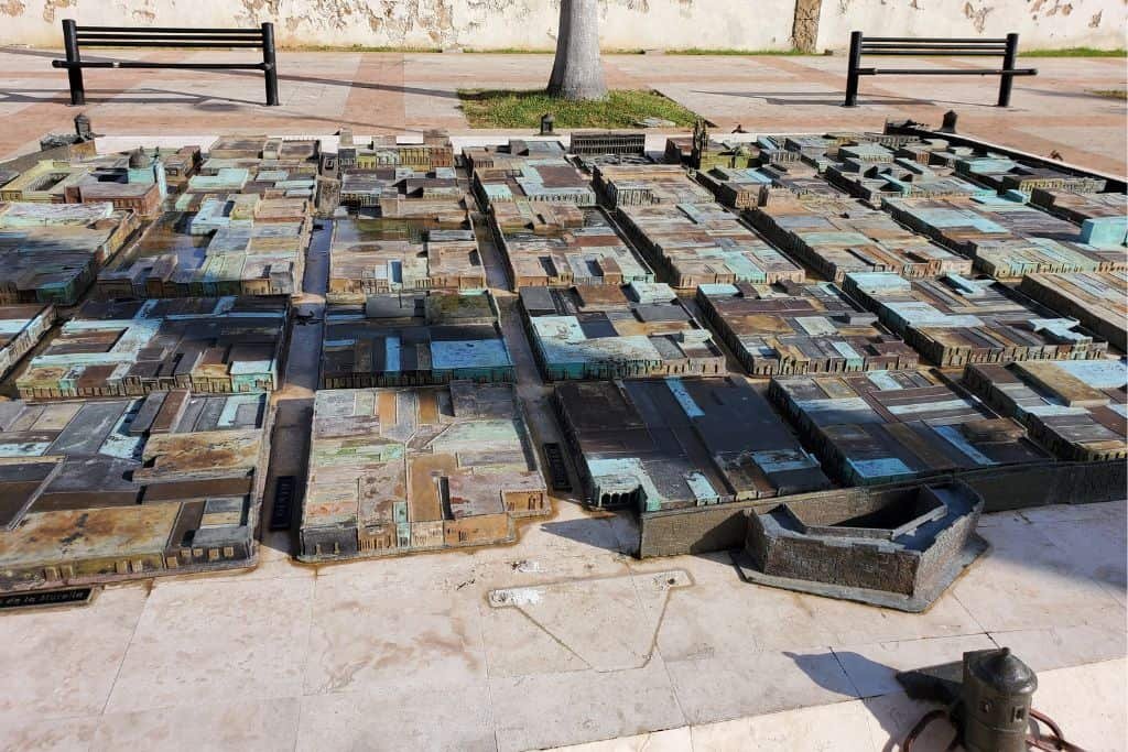 Outdoor bronze 3D model of a city layout, representing historic Campeche, displayed on a stone platform. The model includes buildings and streets in raised relief, with benches and a tree visible in the background.