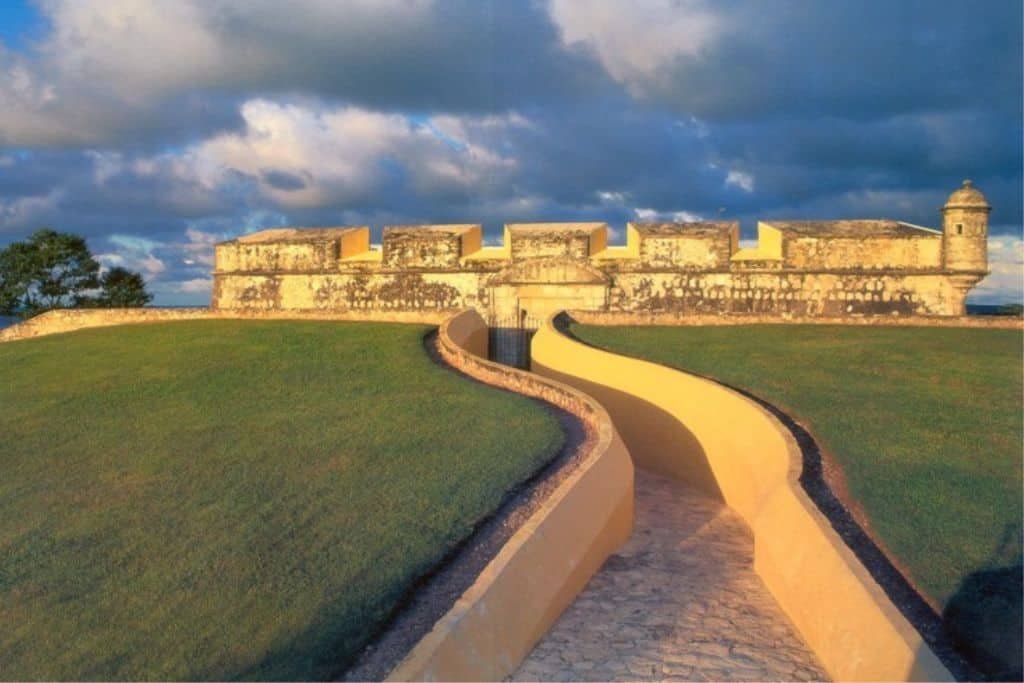 Curving stone path leading up to the grand entrance of Fuerte de San Miguel, a historic yellow-stone fort in Campeche, Mexico. The structure is bathed in warm sunlight under dramatic, cloud-filled skies, sitting atop a grassy hill.