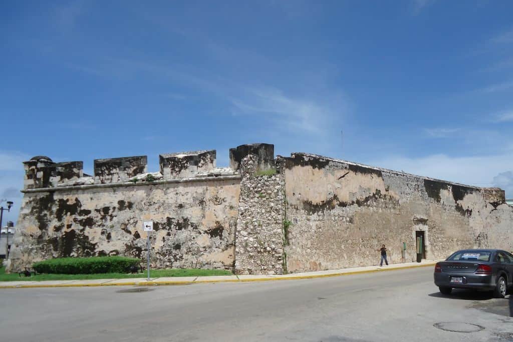 Large stone fortification in Campeche, Mexico, with weathered walls and crumbling battlements, set at the corner of a quiet street. A few parked cars and a pedestrian highlight the scale of the historic structure against a bright blue sky.