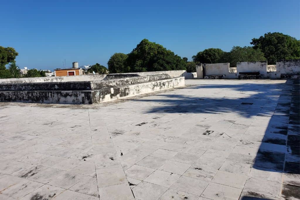 Expansive flat stone surface atop a historic fort or wall in Campeche, Mexico, with low parapets and distant views of trees and buildings under a clear blue sky. The weathered stone is sunlit and mostly open, creating a broad panoramic feel.