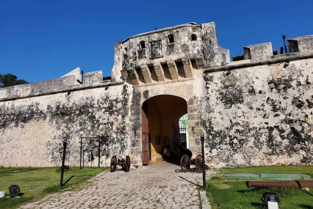 Entrance to a historic fort in Campeche, Mexico, featuring tall weathered stone walls with battlements and a cobblestone path leading through an arched gateway. Old cannons are displayed on either side of the path, set against a backdrop of bright blue sky.
