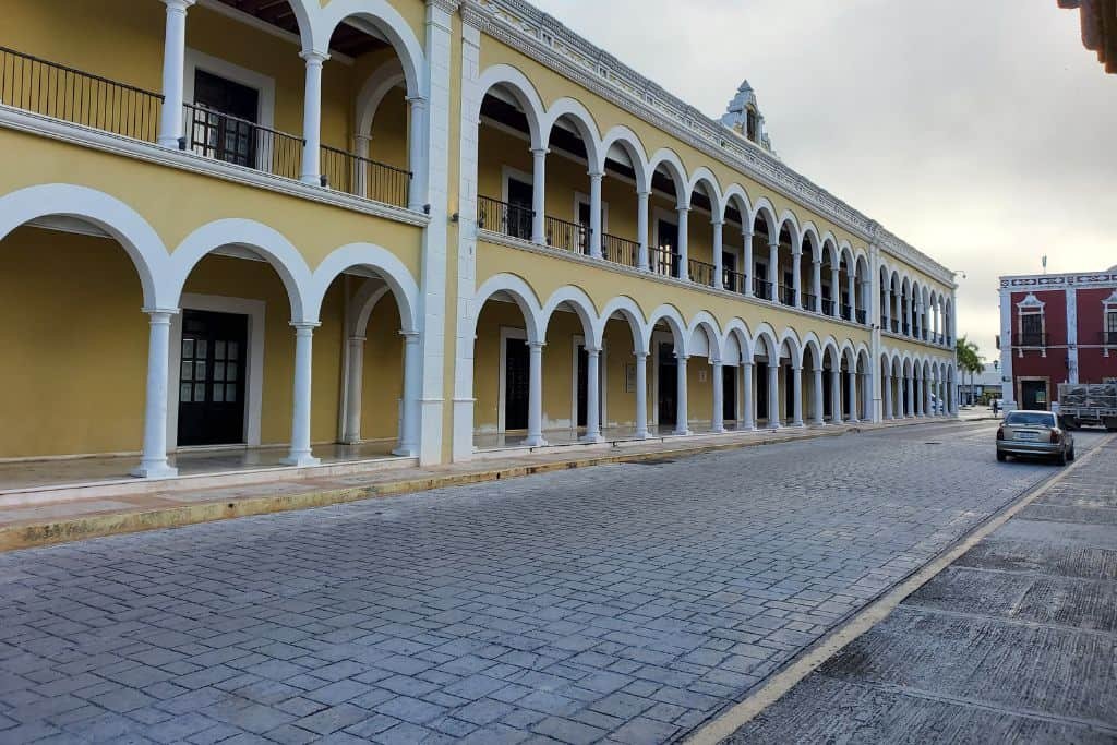 Colonial-style yellow building with white arches and two levels of covered walkways along a quiet cobblestone street in Campeche, Mexico. A few parked cars and a red building in the distance add contrast to the early morning or evening light.