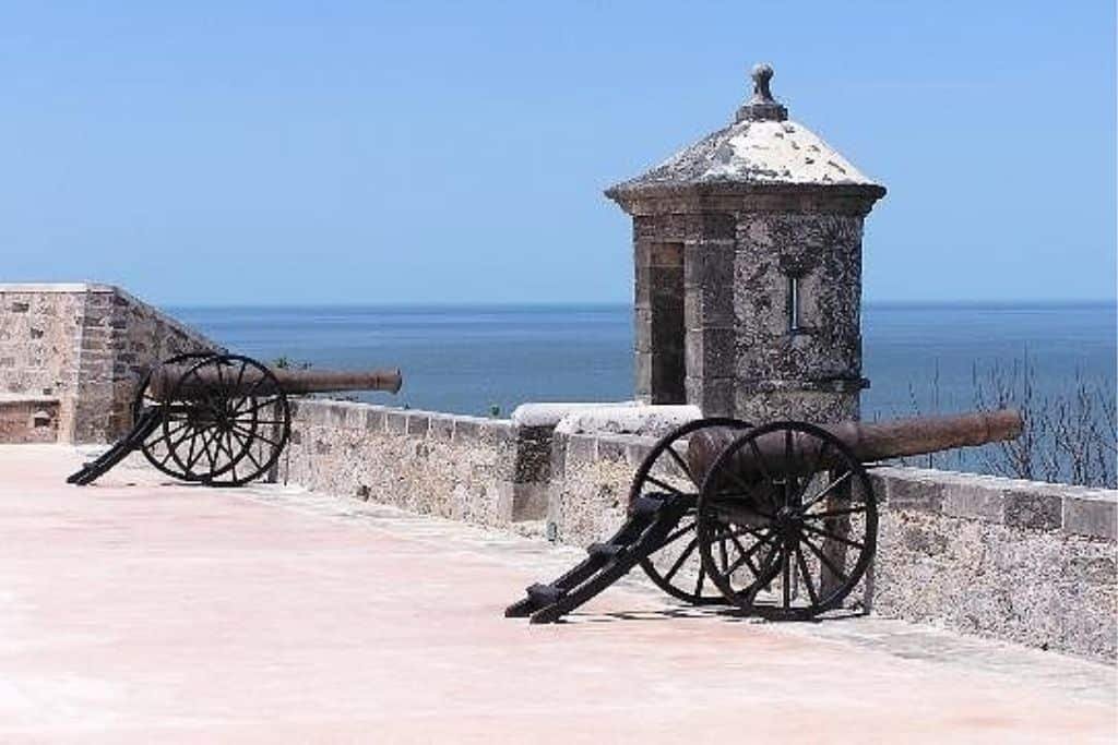 Two historic cannons positioned on a stone fort wall overlooking the Gulf of Mexico in Campeche, Mexico. A small watchtower with a domed roof stands nearby, framed by a clear blue sky and calm sea in the background.