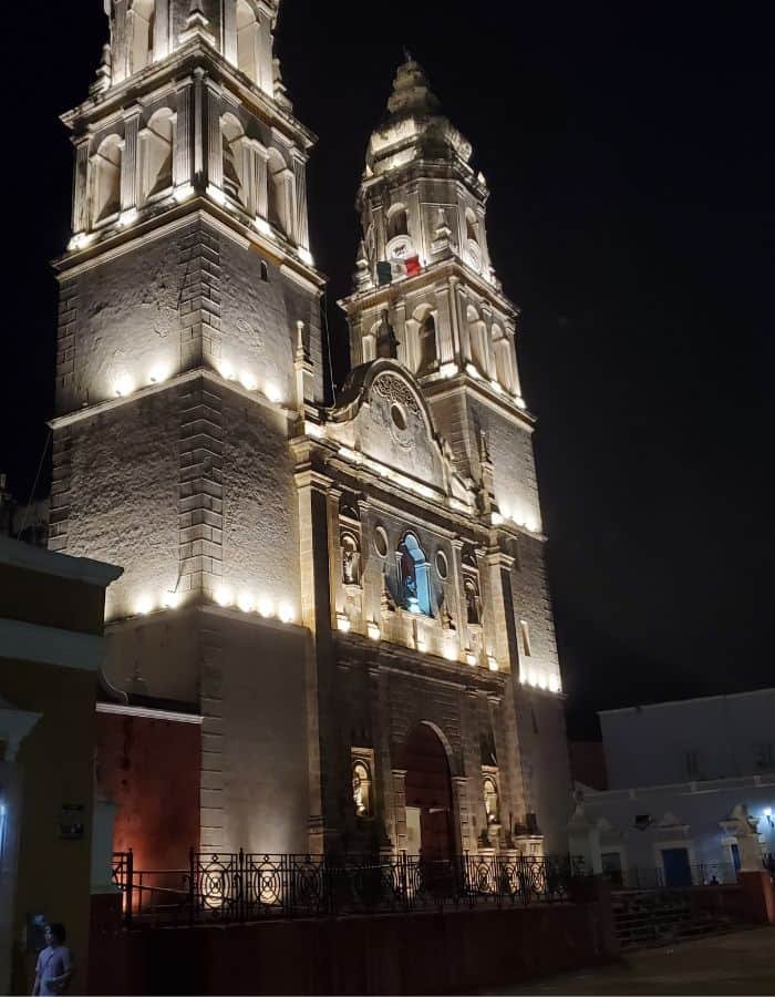 Campeche Cathedral illuminated at night, with its twin bell towers and ornate façade glowing warmly against the dark sky. The grand colonial architecture is highlighted by soft lighting, creating a dramatic and majestic appearance.