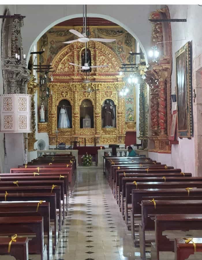 Interior of a historic church in Campeche featuring a golden ornate altar with religious statues in arched niches. The aisle is lined with polished wooden pews, each tied with yellow ribbons, leading to the brightly lit sanctuary.