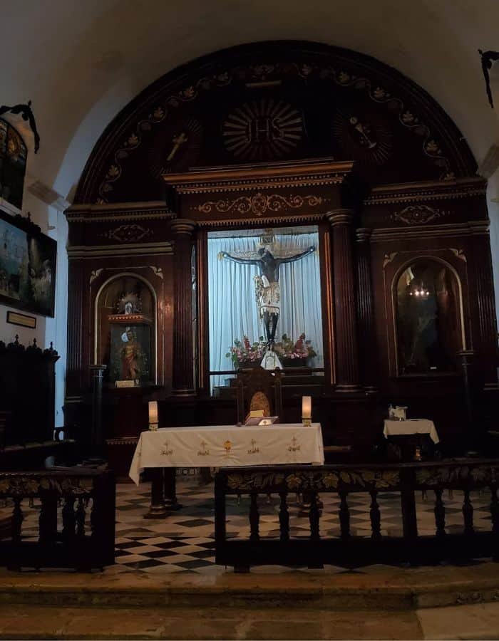 Dimly lit chapel interior in Campeche featuring a prominent crucifix with a black Jesus figure at the center of an ornate wooden altar. The altar is flanked by two statues in niches, with a white-clothed table and candles placed in front.