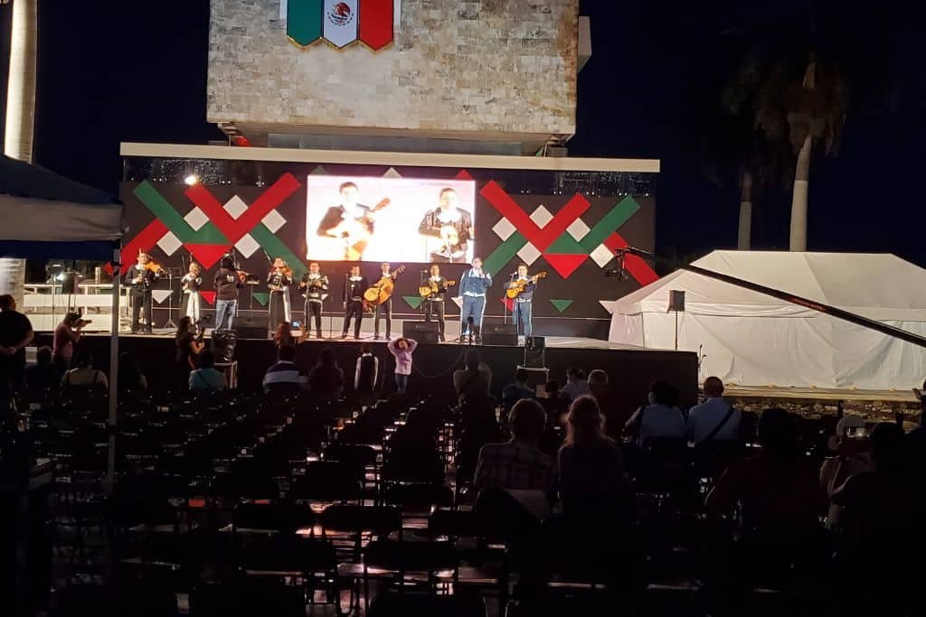Outdoor evening concert in Campeche featuring a mariachi band performing on a stage decorated with red, green, and black patterns and a Mexican flag above. Audience members sit in rows of chairs facing the stage, with a screen behind the performers showing live footage.
