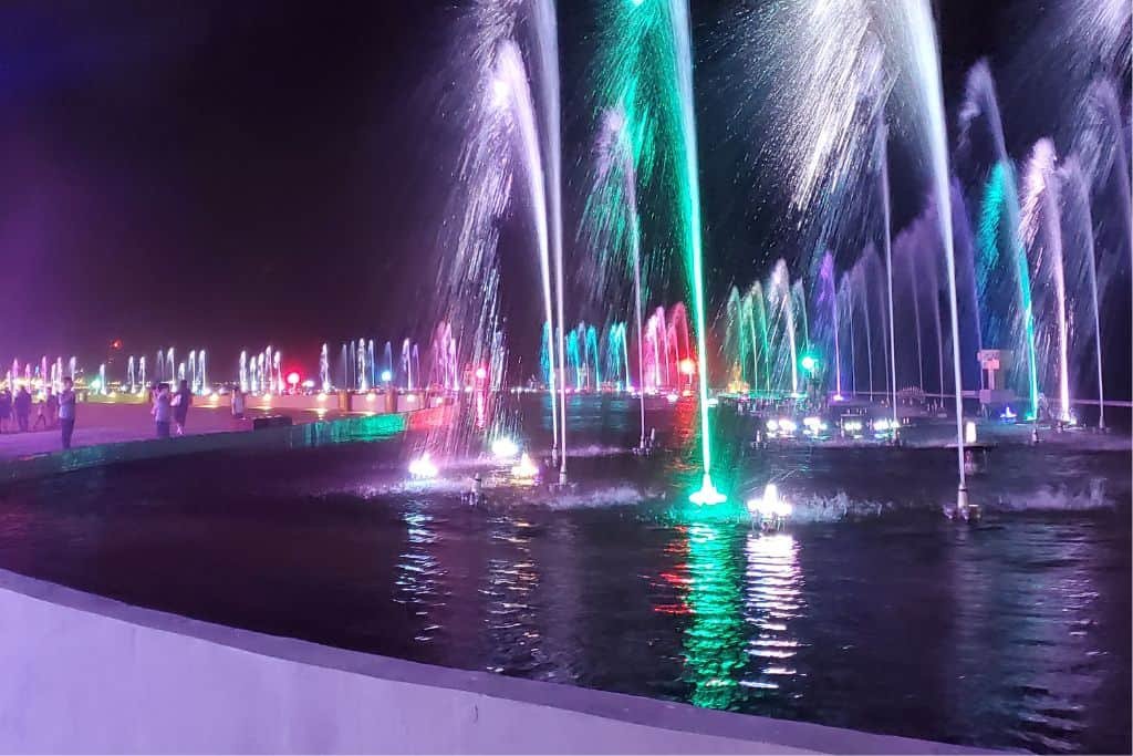 Colorful nighttime fountain display in Campeche with jets of water lit by vibrant green, purple, pink, and blue lights. The illuminated streams reflect on the surface of the large circular pool, creating a festive and lively atmosphere.