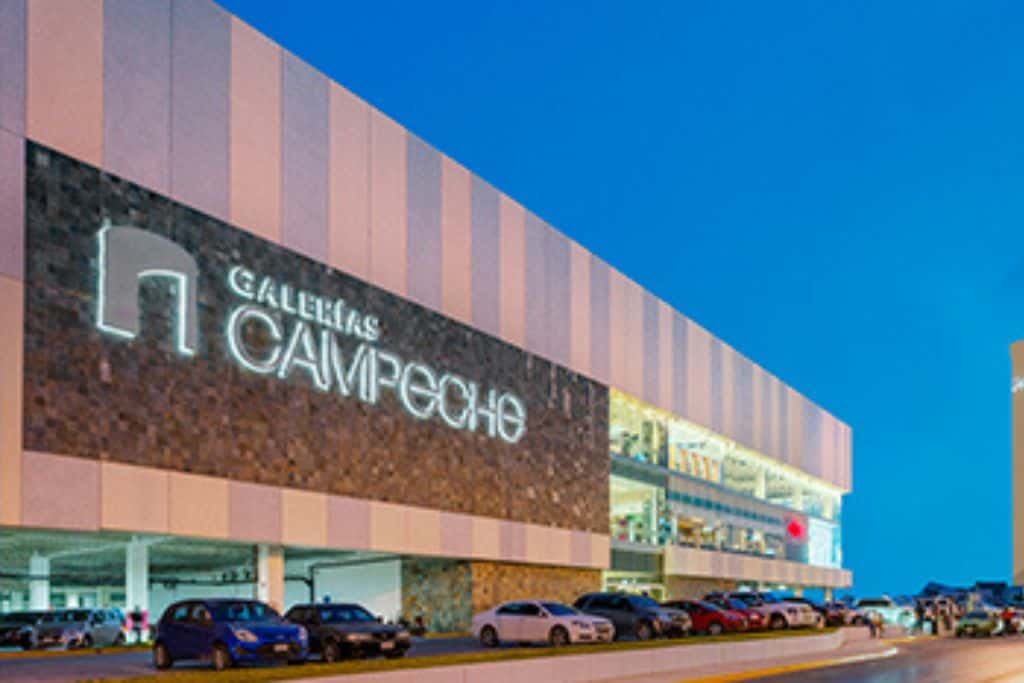 Exterior of Galerías Campeche, a modern shopping mall with a sleek design featuring vertical paneling and a large illuminated sign. Rows of parked cars line the entrance as evening light casts a soft blue glow over the scene.