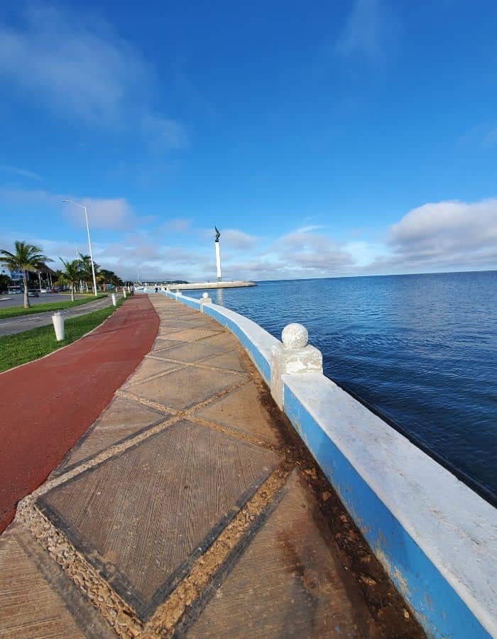 Scenic view of the malecón (seaside promenade) in Campeche, Mexico, with a wide walking and biking path alongside calm blue waters. A white statue on a tall pedestal stands in the distance under a bright sky with scattered clouds.